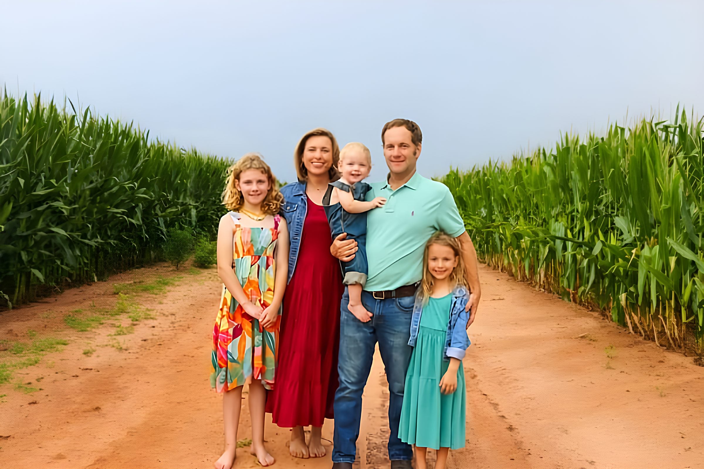 A family of five standing on a red dirt road between two fields of tall corn.