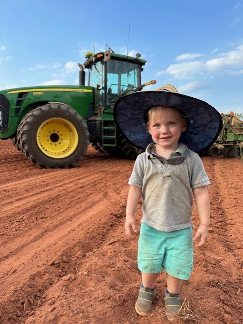 A small child in a wide-brimmed hat, smiling, in a plowed field in front of a green tractor.